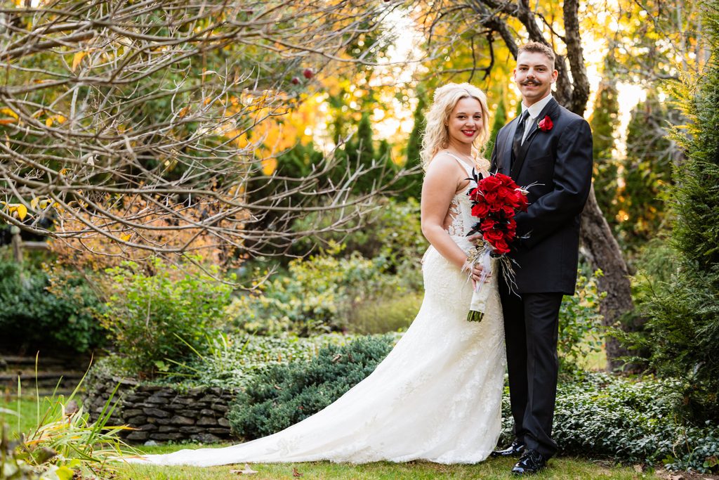 Newlyweds pose beneath golden autumn trees in a garden, with the bride’s long lace train and red rose bouquet on display.