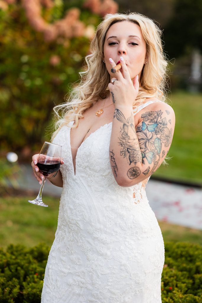 Bride in a lace wedding dress holds a glass of red wine and a cigar during her Lilac Inn wedding in Brandon, Vermont.