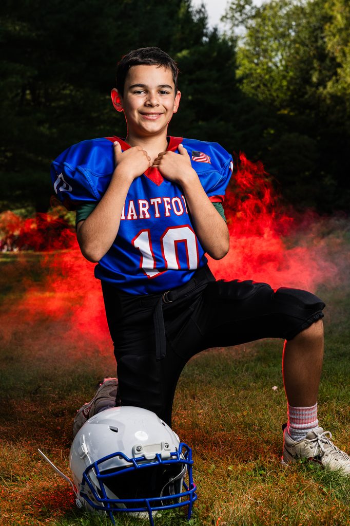 Young football player in a blue number 10 jersey kneels beside a helmet as red smoke drifts through a wooded setting during an outdoor Vermont photo session.