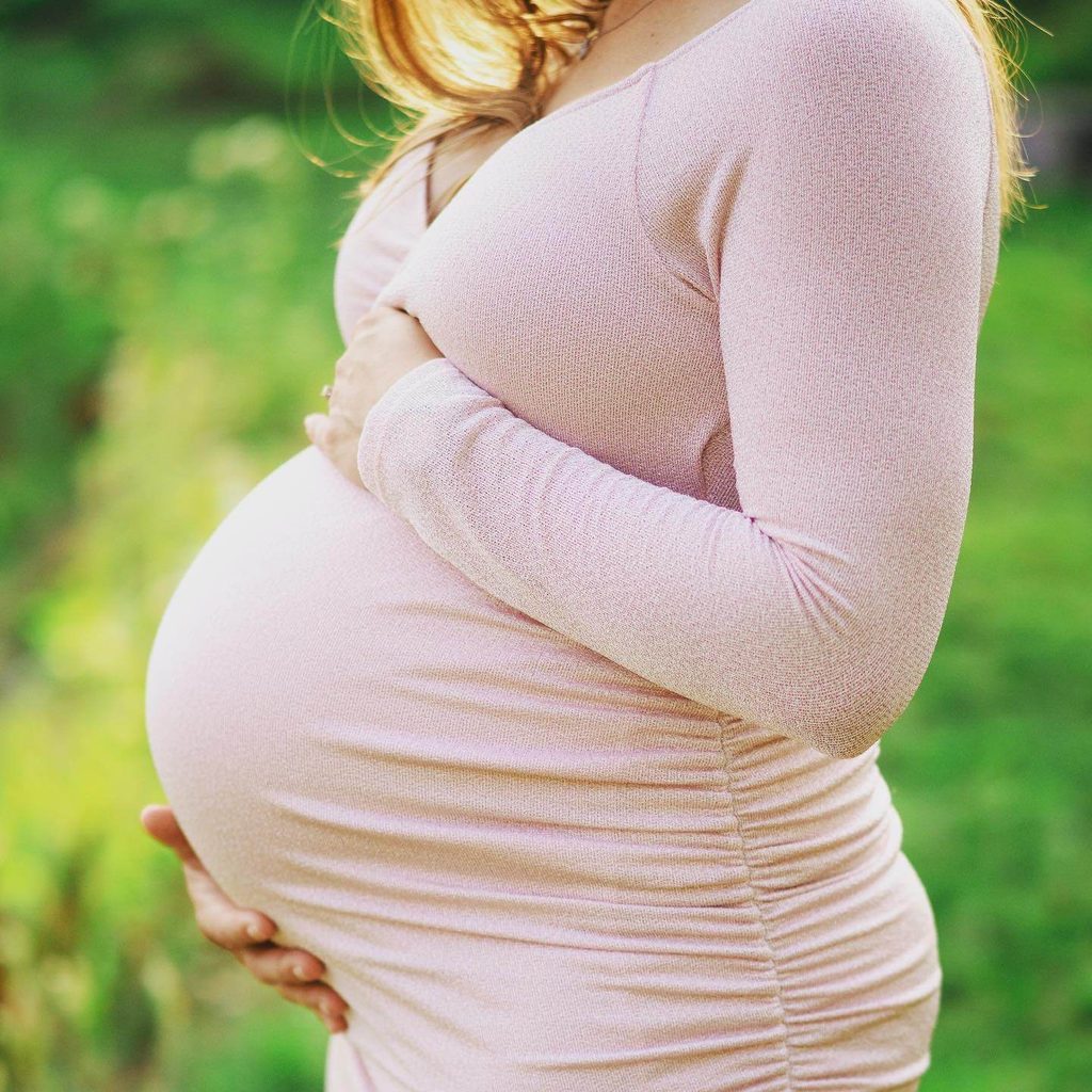 Close-up of an expecting Mom resting their hands on their belly while wearing a soft pink dress outdoors.