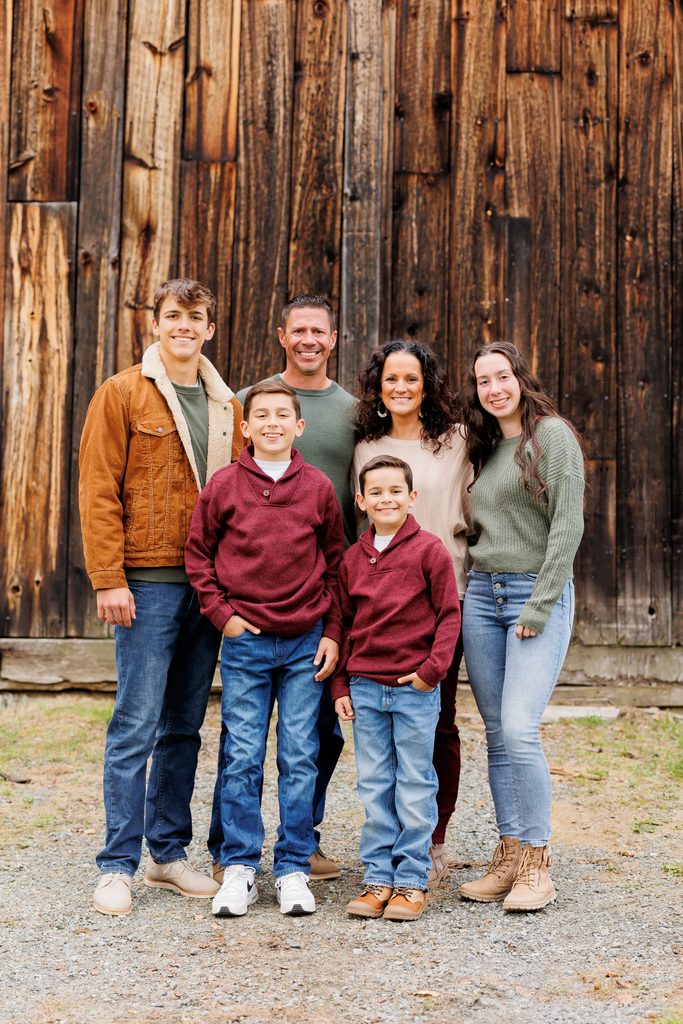 A family of six stands together in front of a weathered wooden barn.