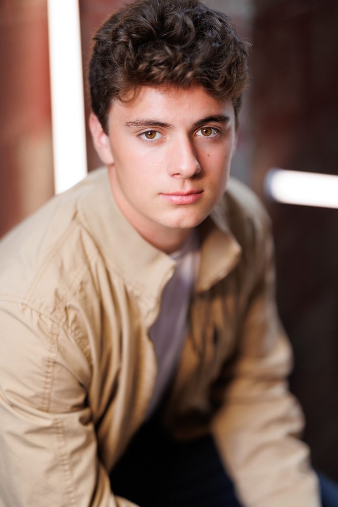 A young man in a tan jacket looks toward the camera in soft indoor light during a Vermont photo session.