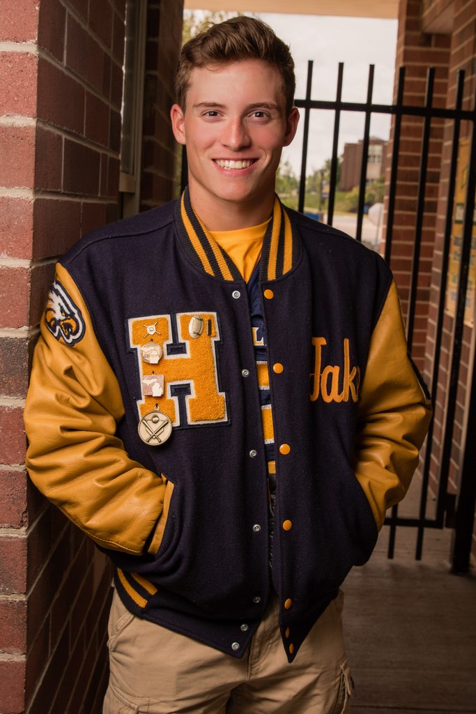 High school senior wearing a letterman jacket in front of a brick wall and iron gate