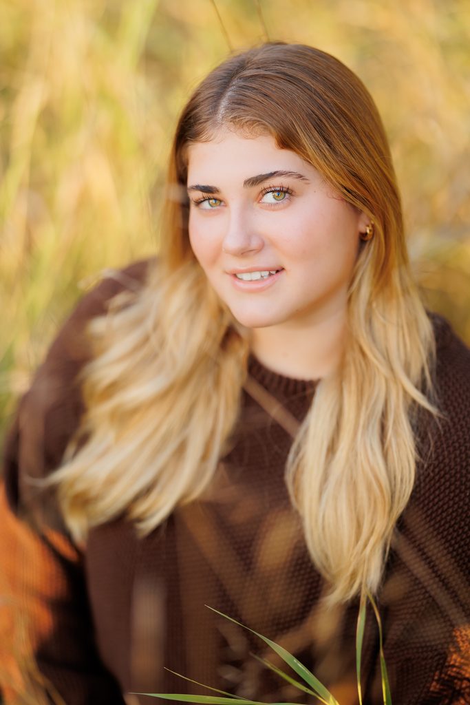 A young woman in a brown sweater smiles among tall grasses in warm golden-hour light.